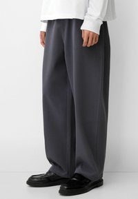Graue, weit geschnittene Hose mit strukturiertem Finish und seitlichen Nähten, kombiniert mit schwarzen Slippern. Baumwollbasierter Stoff, lockere Passform.