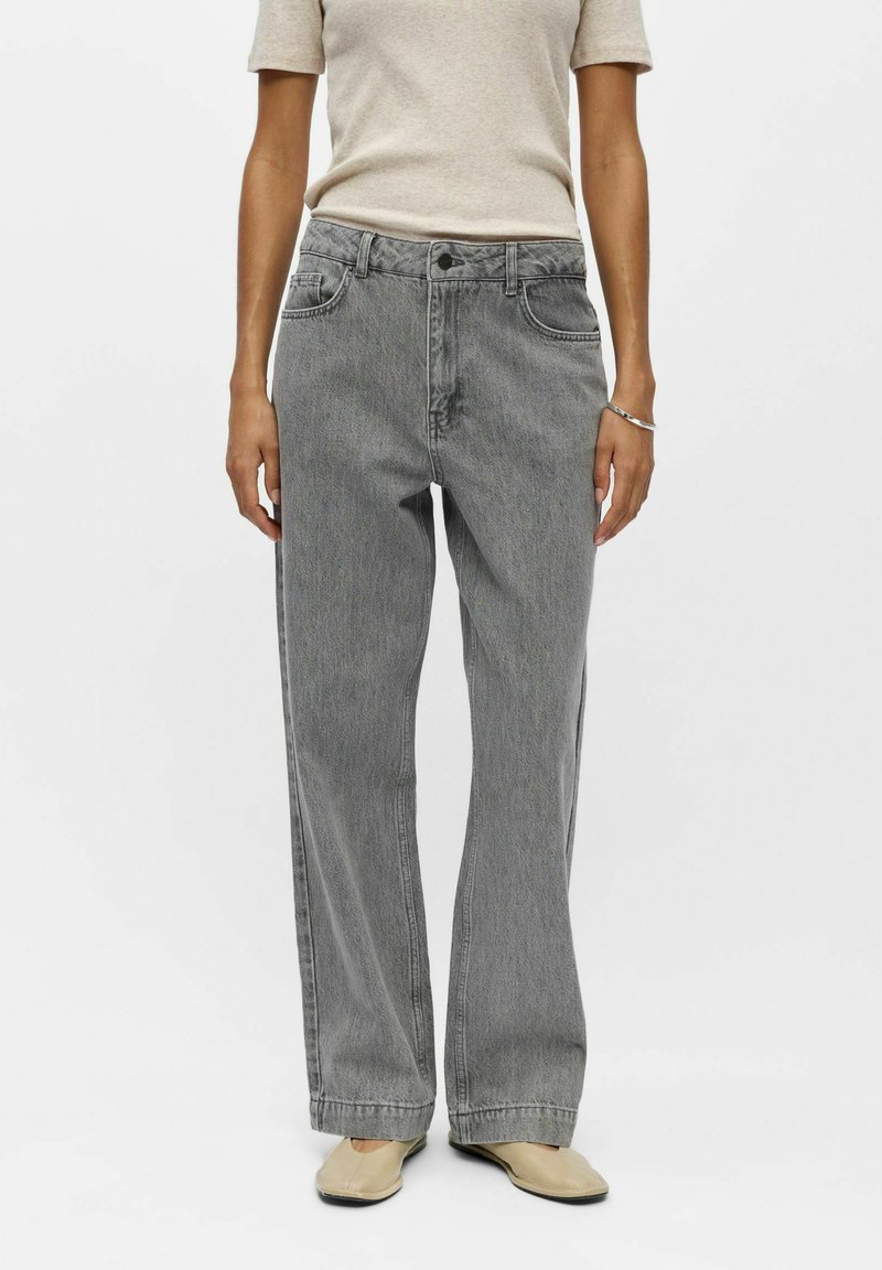Personne portant un jean gris ample taille haute, un haut beige à manches courtes, des chaussures beiges et un bracelet argenté au poignet droit sur fond blanc.