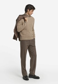 Beżowy sweter z golfem w splot kabelkowy, brązowe dopasowane spodnie oraz czarne mokasyny. Model trzyma brązową kurtkę na jednym ramieniu.