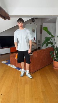 Jeune homme portant un t-shirt gris décontracté, un short noir et des baskets argentées, debout dans un salon moderne avec un canapé marron et une grande plante verte.