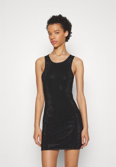 Femme aux cheveux bouclés courts portant une robe mini sans manches en maille noire, se tenant devant un fond blanc uni.