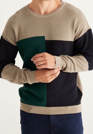 Homme portant un pull beige, vert et noir en blocs de couleurs ajustant la manche, vêtu d’un jean bleu foncé.