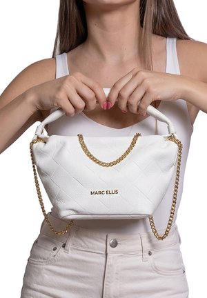 Femme en débardeur blanc et pantalon tient un sac à main blanc Marc Ellis avec une anse en chaîne dorée à hauteur de poitrine.