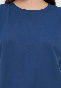 Γυναίκα που φοράει απλό μπλε t-shirt με στρογγυλή λαιμόκοψη, εμφανίζοντας το πάνω μέρος του στήθους και του λαιμού, με μακριά καστανά μαλλιά ορατά.