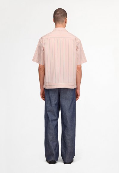 Homme de dos portant une chemise rose clair à manches courtes avec de fines rayures et un pantalon bleu ample sur un fond uni.