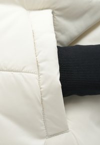 Μπουφάνσαντόν µπεζ χρώματος με ραφές σε σχέδιο καπιτονέ, με μαύρο ριμπ ύφασμα στον μανικό που εκτείνεται από τον καρπό. Μαλακή, λεία υφή.