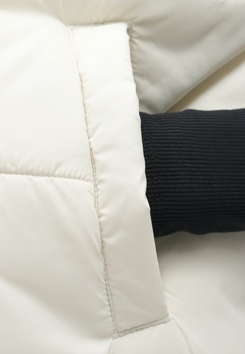 Cremefarbene Steppjacke mit gesteppten Details, die einen gerippten schwarzen Ärmel vom Bündchen ausgehend aufweist. Weiche, glatte Textur.