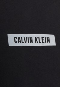 Camiseta de tela negra con una textura suave que cuenta con una etiqueta rectangular blanca con la inscripción "CALVIN KLEIN" en letras negras en negrita.