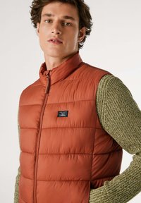 Gilet imbottito in arancione ruggine con colletto alto e chiusura con zip, dotato di maniche in maglia verde testurizzata, logo del marchio sul lato sinistro del petto.