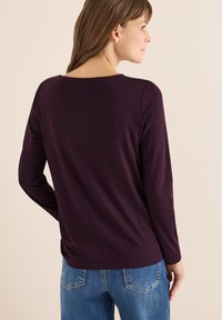 Long-sleeve purple top made of soft fabric. Features a rounded neckline and a relaxed fit, paired with blue jeans and a smooth texture.