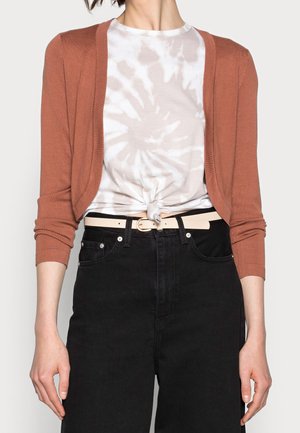 Femme portant un cardigan court couleur rouille sur un tee-shirt blanc noué tie-dye, un jean noir taille haute et une ceinture beige clair.