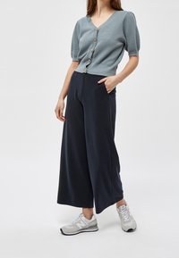 Světle modrý pletený cardigan s krátkými rukávy a dřevěnými knoflíky, kombinovaný s tmavými širokými kalhotami a šedými teniskami. Jednoduchý a ležérní outfit.