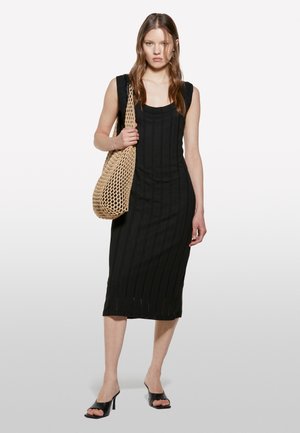 Schwarzes, geripptes Midikleid mit taillierter Silhouette und seitlichen Schlitzen, kombiniert mit einer gewebten Tasche in Erdtönen und offenen schwarzen Absatzsandalen.