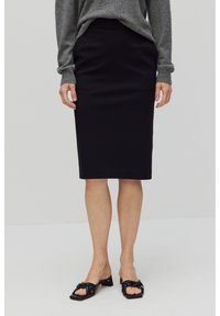 Jupe noire longueur genou en tissu lisse, présentant une silhouette ajustée. Associée à des chaussures à enfiler noires nouées avec des petits talons.