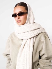 Egy nő fekete tojásdad napszemüveget visel, vastag krémszínű kötött sálat tekerve a fejére és a nyakára, bézs kabátban.