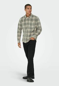 Siva in zelena šahovnica s dolgimi rokavi s zapiranjem na gumbe, v paru z črnimi hlačami in črnimi škornji. Model stoji na enobarvni podlagi.