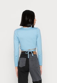 Top cropped de manga longa azul claro com um decote redondo, apresentando um detalhe de laço nas costas. Usado com jeans pretos bicolor de cintura alta.