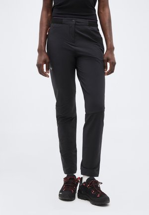 Άτομο που φοράει μαύρο παντελόνι για υπαίθριες δραστηριότητες και μαύρα ορειβατικά παπούτσια με κόκκινες κορδόνια, στεκόμενο μπροστά σε απλό φωτεινό φόντο.