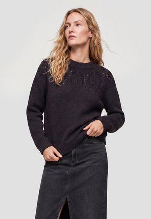 Kvinde med langt blondt hår iført en sort striktrøje og et mørkt denimsnørklæde med en frontslids, der kigger let til siden.