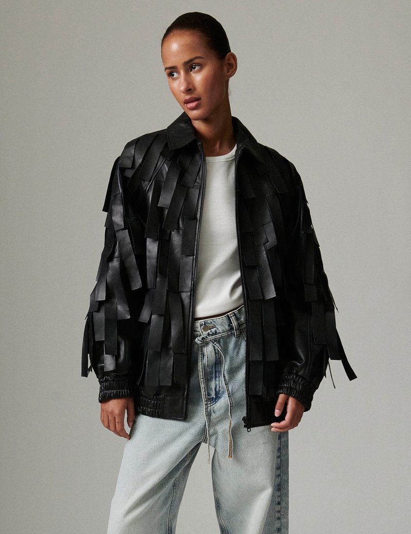 Μαύρο δερμάτινο μπουφάν bomber με υφασμένη φράντζα, κλείσιμο με φερμουάρ και ριμπ μανσέτες. Φορεμένο πάνω από άσπρο τοπ και τζιν σε ανοιχτή απόχρωση.