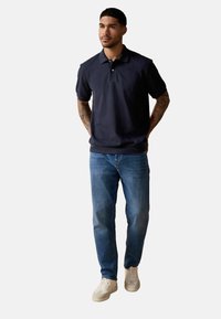 Marineblå polo-shirt med korte ærmer, knappet krave, parret med lyseblå jeans og hvide sneakers, stående foran en lys baggrund.