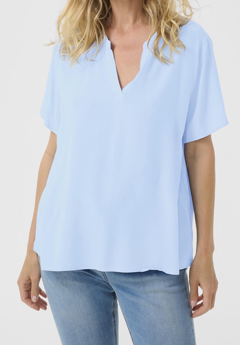 Femme portant un chemisier bleu clair à manches courtes et col en V ainsi qu’un jean bleu clair, debout, les bras détendus le long du corps.