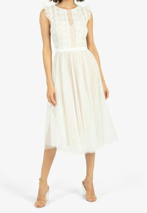 Frau trägt ärmelloses weißes Spitzenkleid mit gerafften Schultern und Tüllrock, kombiniert mit Sandalen mit klarem Absatz und Knöchelriemen.
