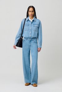 Camicia di denim blu chiaro con due tasche frontali, abbinata a jeans a gamba larga coordinati. Presenta una camicia bianca con colletto sotto e scarpe marroni.