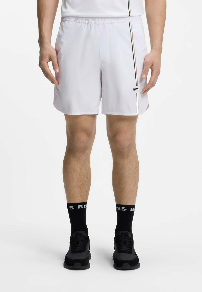 Homme portant un short de sport blanc avec une bande verticale et le logo "BOSS", des chaussettes noires "BOSS" et des baskets noires, debout devant un fond blanc.