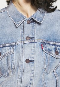 Ljusblå jeansjacka med knappstängning, spetsig krage och två bröstfickor. Har kontrastsömmar och en liten röd logotypetikett.