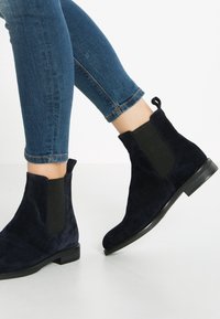 Botines de ante azul marino con paneles elásticos en los laterales, suelas planas y tiradores. Combinados con unos vaqueros cortos de color azul oscuro.