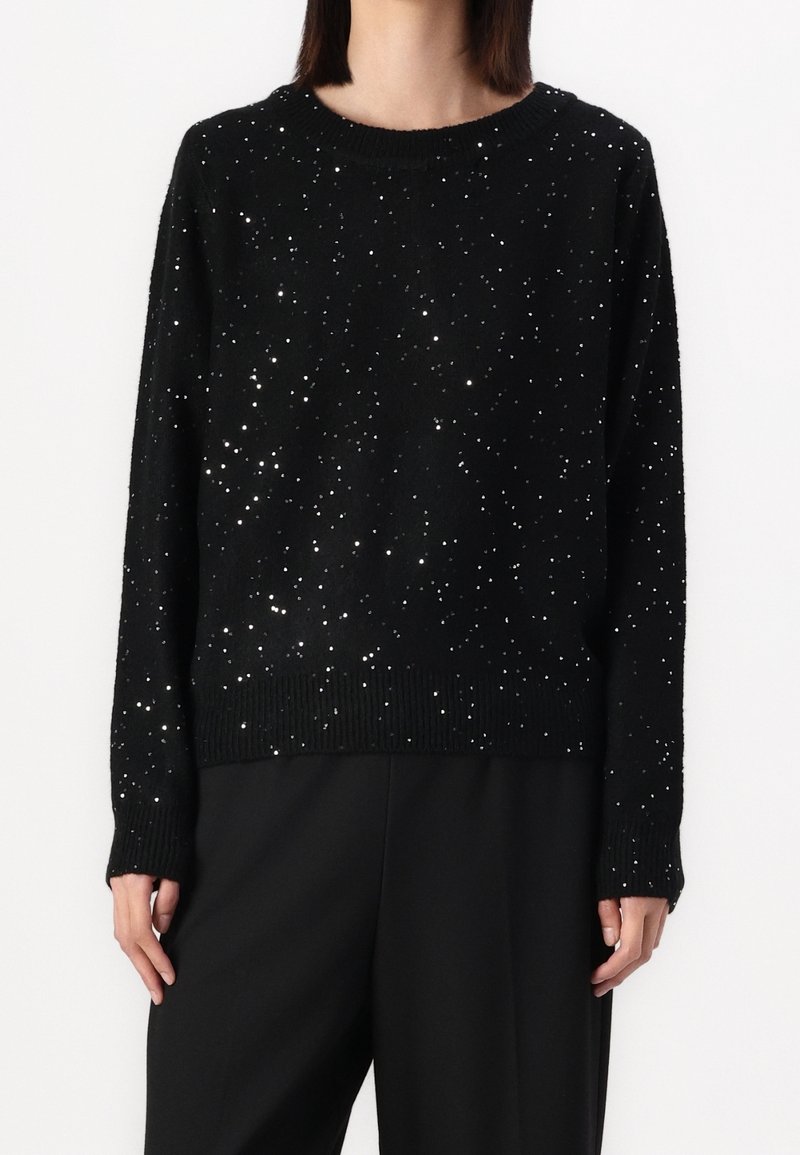 Personne portant un pull noir à manches longues avec de petites ornements brillants et un pantalon noir, debout devant un fond uni.
