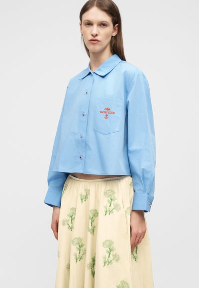 Γυναίκα με ανοιχτό γαλάζιο πουκάμισο με κέντημα "Yacht Club" και κρεμ φούστα με πράσινα λουλουδένια σχέδια, που στέκεται μπροστά σε λευκό φόντο.