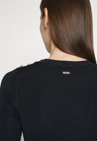 Femme aux longs cheveux bruns portant un pull côtelé noir avec des boutons argentés sur l'épaule et un petit logo "BOSS" au dos du col.