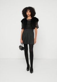 Colete de pele falsa preta sobre uma blusa preta transparente, combinado com uma saia plissada cinza escuro. A modelo usa collants pretos e sapatos de salto alto, segurando uma pequena mala.