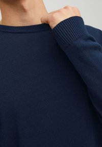 Mężczyzna trzyma granatowy prążkowany sweter z okrągłym dekoltem za kołnierz, pokazując fakturę dzianiny i dopasowany mankiet rękawa na jasnym tle.