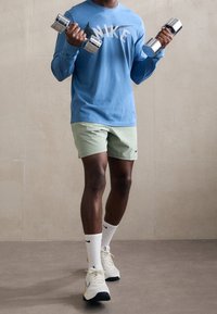 Osoba ve modrém tričku s dlouhým rukávem Nike, světle zelených šortkách, bílých ponožkách Nike a teniskách, která vevnitř zvedá dvě stříbrné činky.