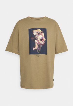 Duży beżowy T-shirt z nadrukiem abstrakcyjnego portretu bocznego twarzy osoby w odcieniach beżu, fioletu i czerni na ciemnym, prostokątnym tle.