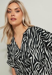 Blusa a righe zebrate in bianco e nero con scollatura a V e maniche corte. Caratterizzata da una tasca sul petto e un tessuto leggero.