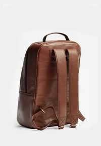 Ruskea nahkainen reppu, jossa on sileä pinta, varustettuna pehmustetuilla olkahihnoilla ja ylhäällä olevalla kahvalla. Minimalistinen muotoilu, jossa on hillityt ompeluyksityiskohdat.