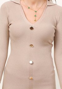 Robe beige à côtes avec un décolleté en V, des boutons au centre et un col. Présente un collier doré superposé avec des accents verts.