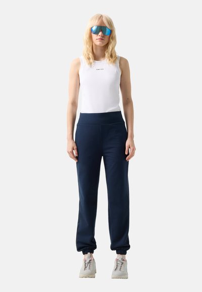 Blonde Frau mit blauen verspiegelten Sonnenbrillen, weißem ärmellosem Top mit dem Text "FIRE + ICE", marineblauen Jogginghosen und weißen Turnschuhen, stehend.