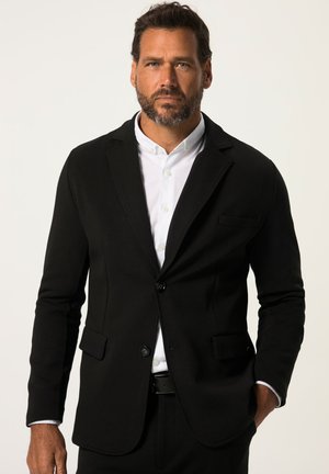 Blazer negro a medida con dos bolsillos frontales y cierre de un solo botón, llevado sobre una camisa blanca con cuello, que presenta una textura suave.