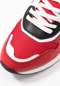 Baskets rouges en mesh et cuir, avec des lacets blancs, un accent noir près de la pointe et une surface texturée.