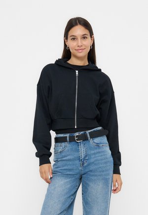 Ung kvinde iført en sort hættetrøje med lynlås og lyseblå jeans med sort bælte, stående og smilende mod en hvid baggrund.