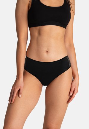 Femme portant des sous-vêtements noirs sans coutures et un soutien-gorge de sport noir assorti, debout avec les bras détendus le long du corps sur un fond uni.