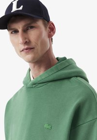Grüner Baumwollhoodie mit einer Vorderseite und einer Kapuze, der ein kleines gesticktes Logo aufweist. getragen mit einer blauen Mütze, die einen weißen Buchstaben zeigt.
