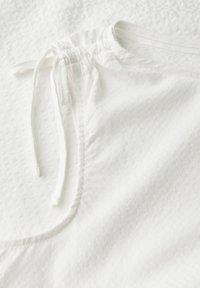 Close-up of a textured white garment featuring a gathered neckline with a tie, set against a white backdrop.