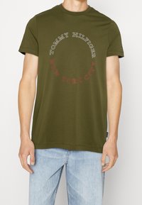 Camiseta de algodón color verde olivo con un gráfico circular que presenta "TOMMY HILFIGER" y "NEW YORK CITY" impresos en blanco y rojo. Manga corta.