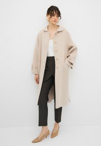 Cappotto in lana beige con una vestibilità rilassata, ampio colletto e quattro bottoni. Abbinato a pantaloni scuri a righe sottili e décolleté beige a punta.
